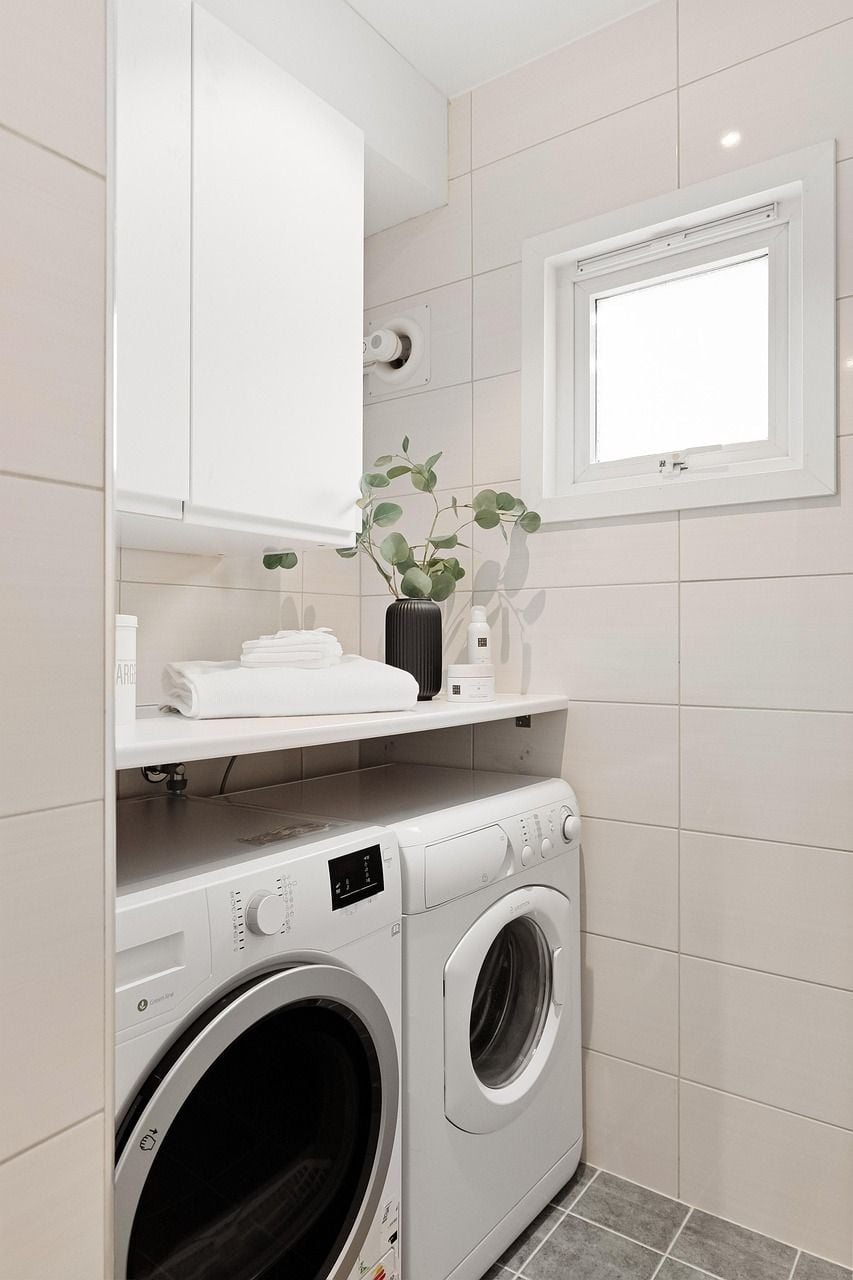 Lavanderia moderna com máquina de lavar instalada sob prateleira com toalhas dobradas e planta