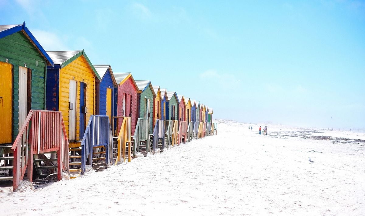 Fileira de casas de praia coloridas na areia, com céu azul ao fundo
