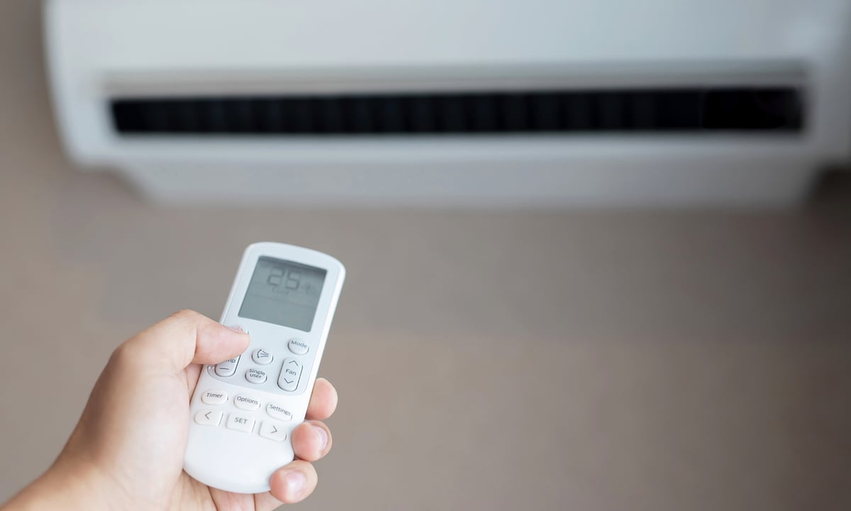 Mão segurando o controle remoto de um ar-condicionado com o aparelho ligado ao fundo